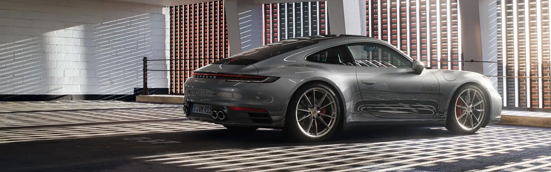 Rear three-quarter view of a grey Porsche 911 parked in a garage with sunlight streaming in.