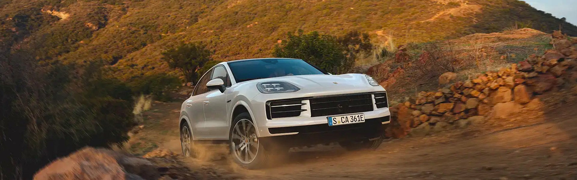 A white Porsche Cayenne SUV drives on a dusty off-road trail in a rocky, mountainous landscape.