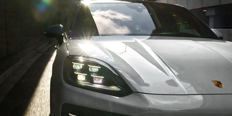 Close-up of a silver Porsche Cayenne's front, with headlights illuminated, sunlight glinting off its hood.