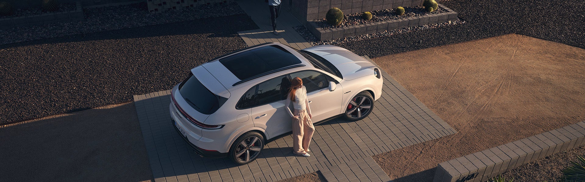 Overhead shot of a woman with red hair leaning against a white SUV parked on a paved driveway.