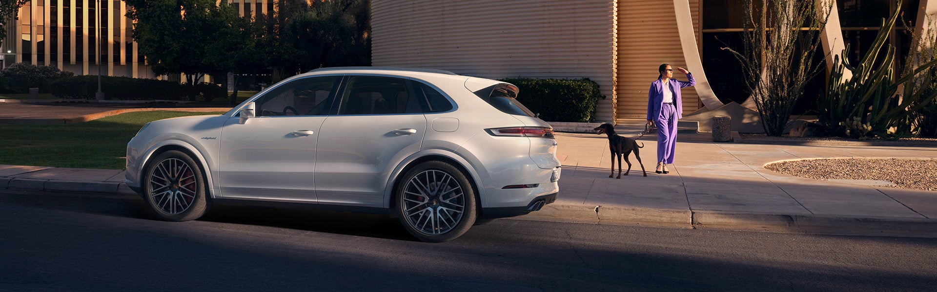 A white Porsche SUV is parked on a street with a woman and black dog on the right.