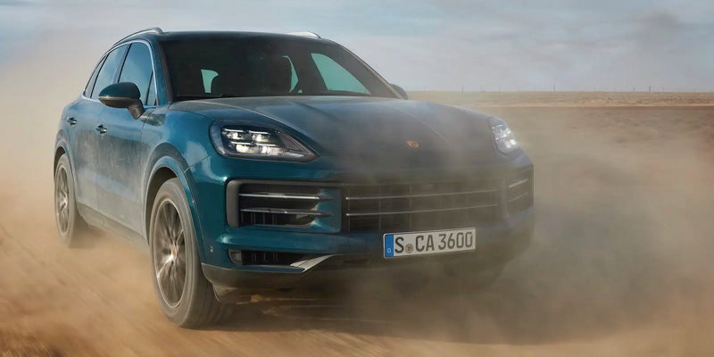 A teal Porsche Cayenne SUV driving on a dirt road, kicking up a large cloud of dust.