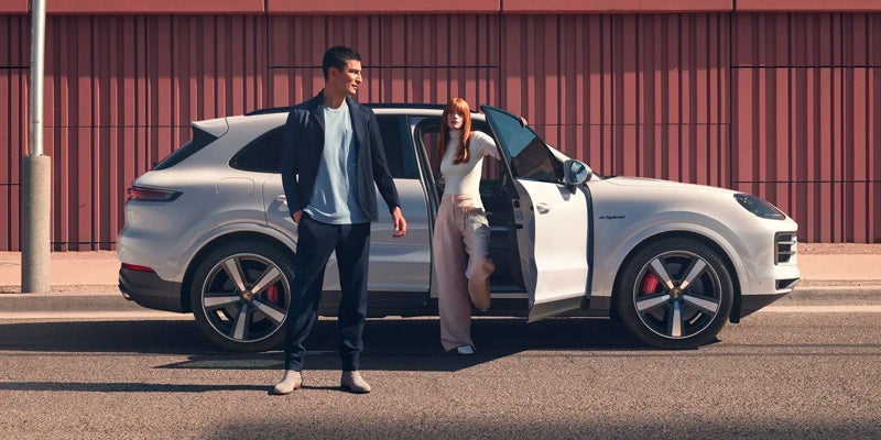Man and woman exiting a white Porsche Cayenne SUV on a sunny day with a red corrugated wall in the background.