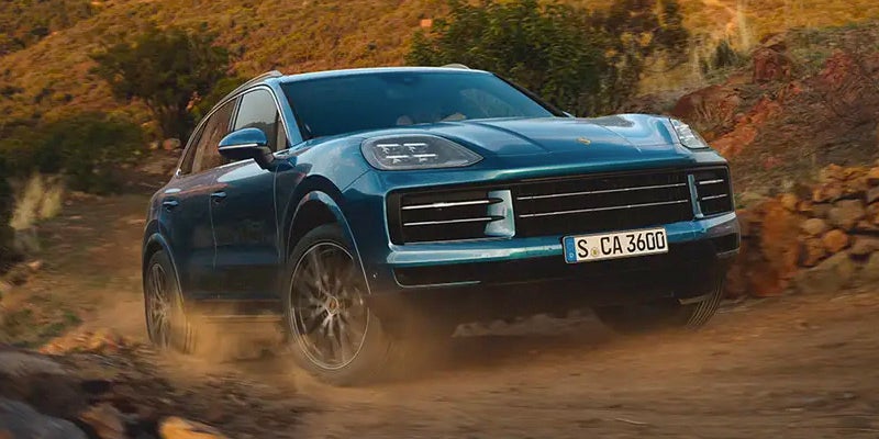 A teal Porsche Cayenne driving up a dusty dirt road, sand kicking up from its tires.