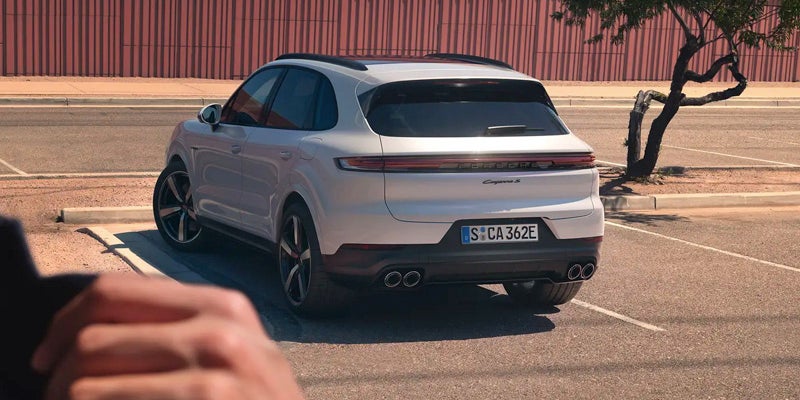 Rear view of a white Porsche Cayenne S parked in a lot, with partial view of a person's hands in the foreground.