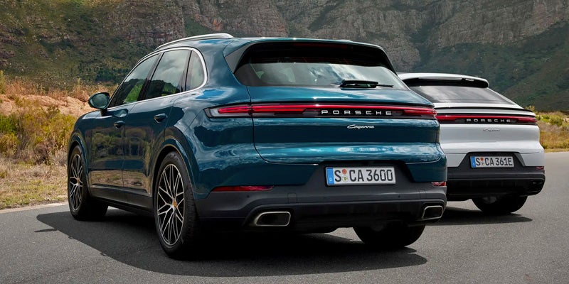 Rear view of a teal Porsche Cayenne parked in front of a white Porsche Cayenne, with mountains in the background.