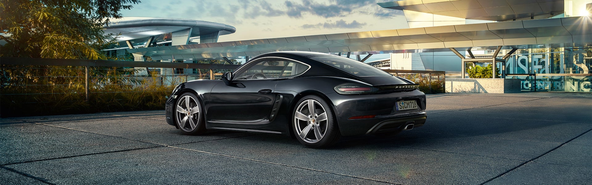 Black Porsche 718 Cayman parked on a paved lot beside an abstract building, sunlight reflecting off the car.