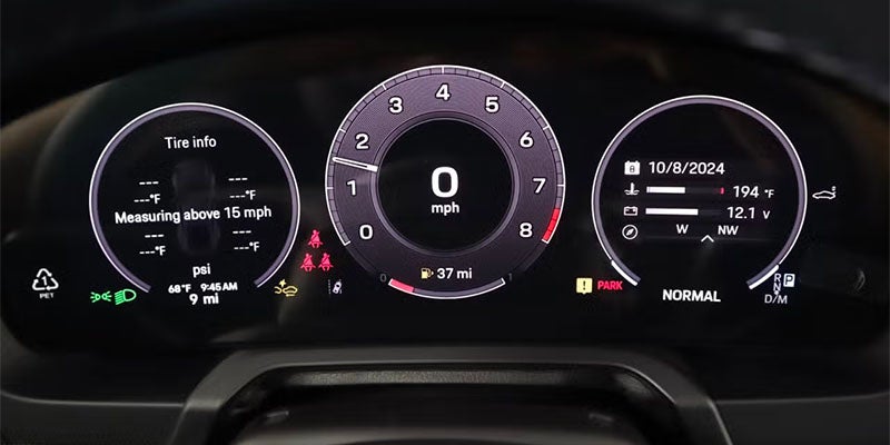 A car's digital dashboard showing tire info, a speedometer at 0 mph, and system info including date and temperature.