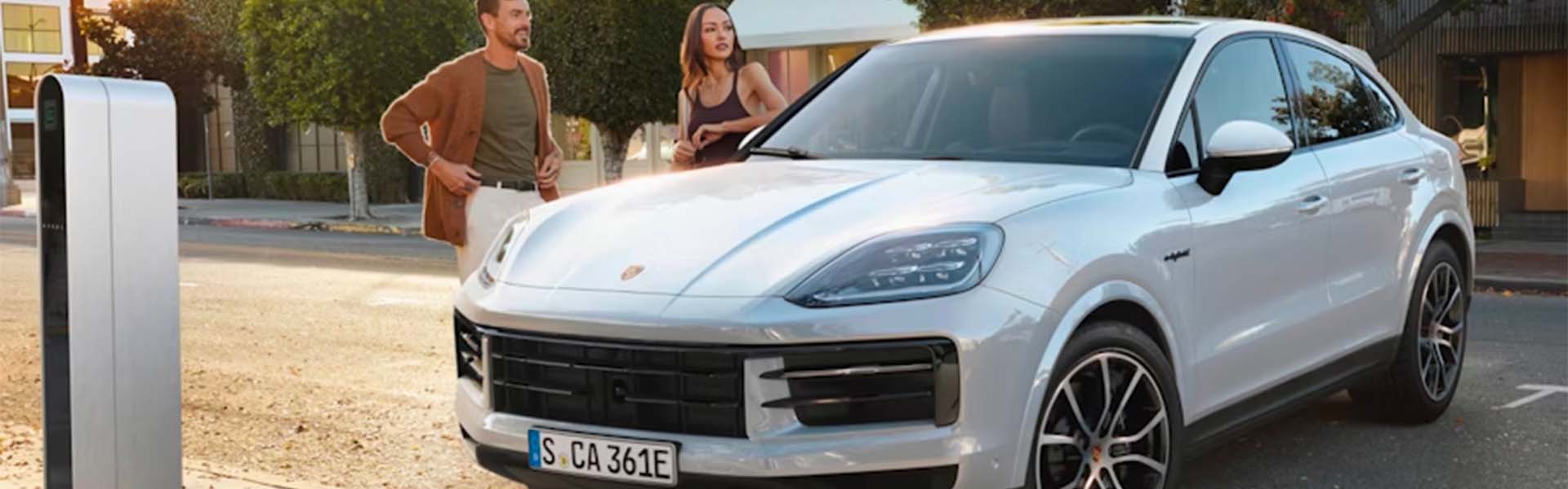 A couple stands next to a white Porsche SUV at an outdoor electric vehicle charging station.