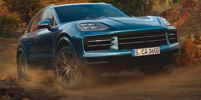 A teal Porsche Cayenne SUV driving fast on a dirt road, kicking up dust.