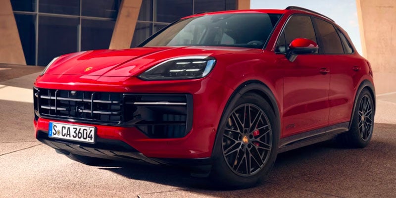 A front 3/4 view of a shiny red Porsche Cayenne GTS, parked outdoors with a beige architectural background.