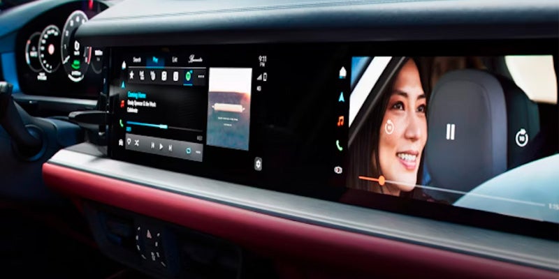 Wide dashboard screen in a car showing a navigation map, music player, and a woman watching a video.