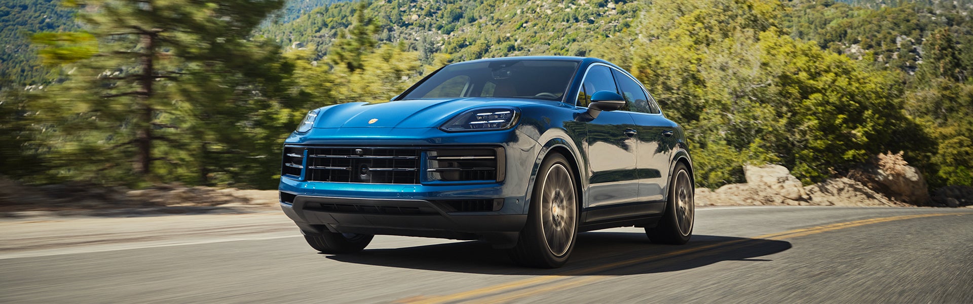 A blue Porsche Cayenne coupe drives quickly along a winding mountain road with green trees in the background.