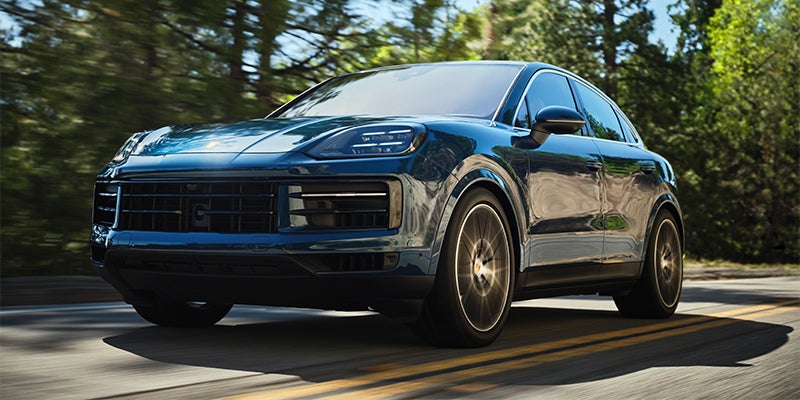 A shiny dark blue Porsche SUV driving on a road lined with trees, captured at a low angle.