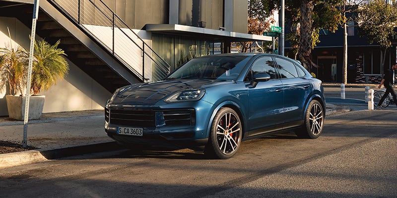A dark blue Porsche Cayenne Coupe parked on a city street with a building and stairs in the background.