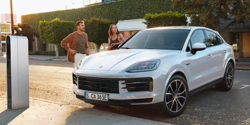 A light gray Porsche Cayenne Coupe parked on a street with a couple standing nearby, an EV charger to the left.