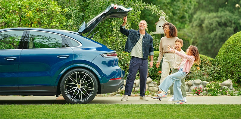 A family of four smiles beside their blue SUV with the trunk open, surrounded by lush greenery.