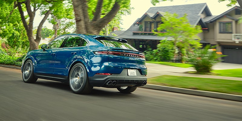 A blue Porsche Cayenne coupe drives away on a suburban street lined with trees and houses.