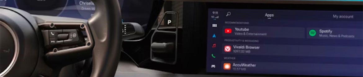 Car interior showing steering wheel, gear selector in P, and a screen displaying apps like YouTube, Spotify, and Vivaldi.