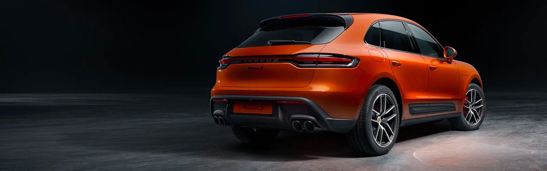 Rear view of a brilliant orange Porsche Macan S on a dark background, showcasing its sleek design.