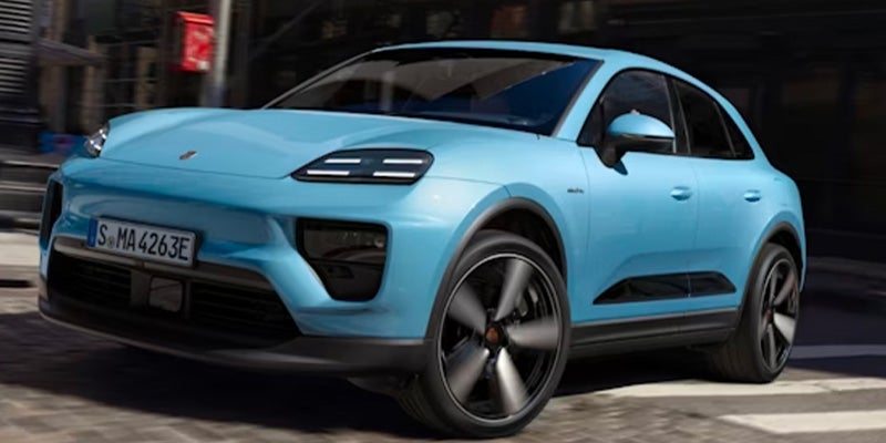 Light blue electric Porsche Macan SUV driving on a road, a building visible in the background.