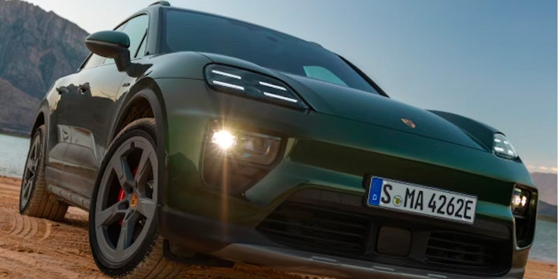 Front view of a dark green Porsche Macan EV on a dirt road, facing slightly left, with mountains and water behind.
