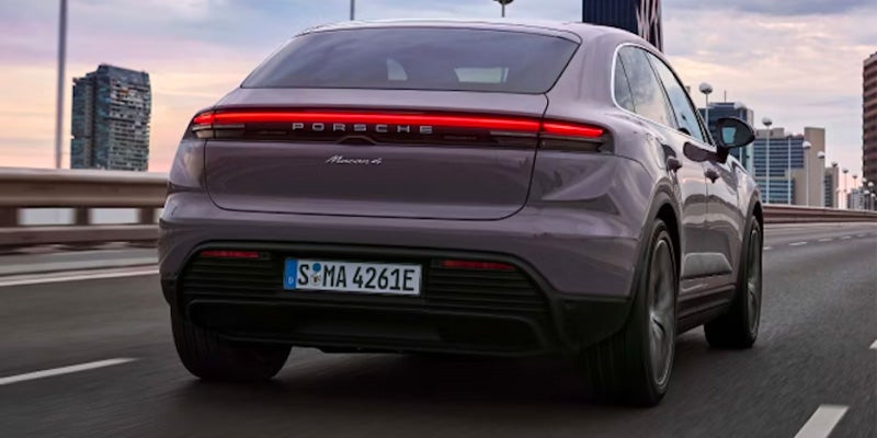 Rear view of a dark gray Porsche Macan 4 driving on a highway with city buildings in the background.