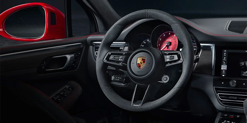 Interior of a black Porsche Macan, showing the steering wheel, red side mirror, speedometer, and infotainment screen.