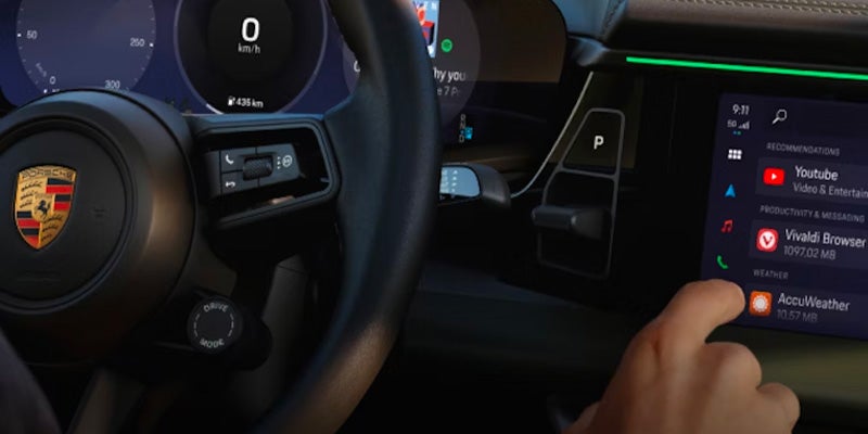 Hand touching infotainment screen showing apps like YouTube and AccuWeather in a Porsche with steering wheel visible.
