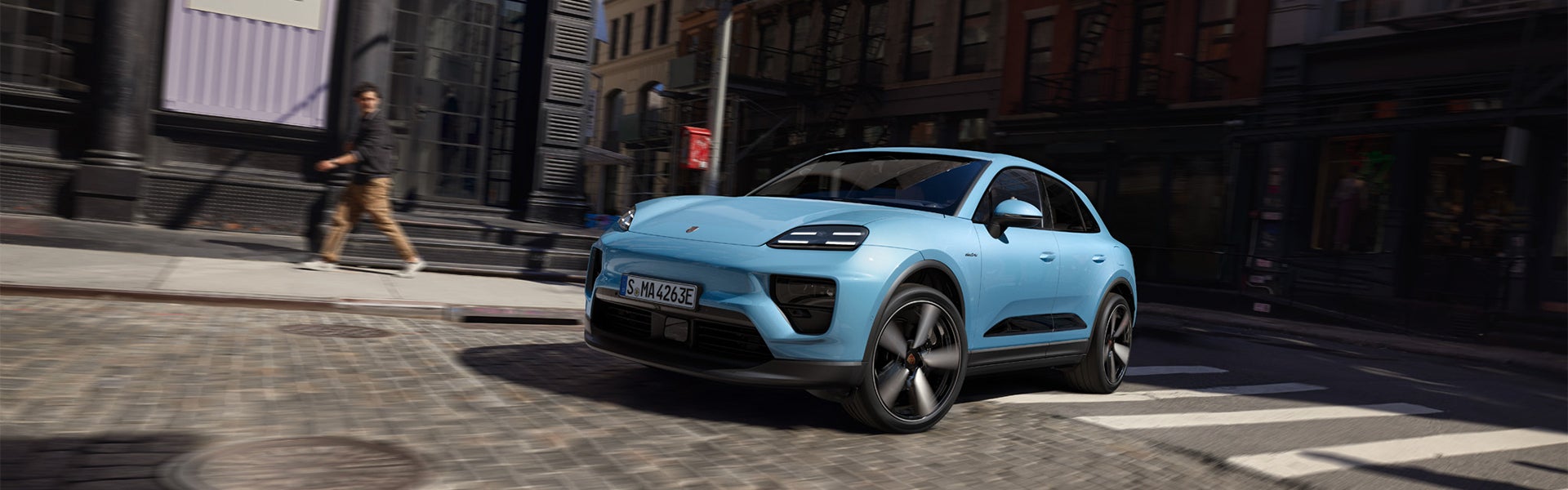 Light blue Porsche Macan driving on a cobblestone street with a blurred man walking on the sidewalk.