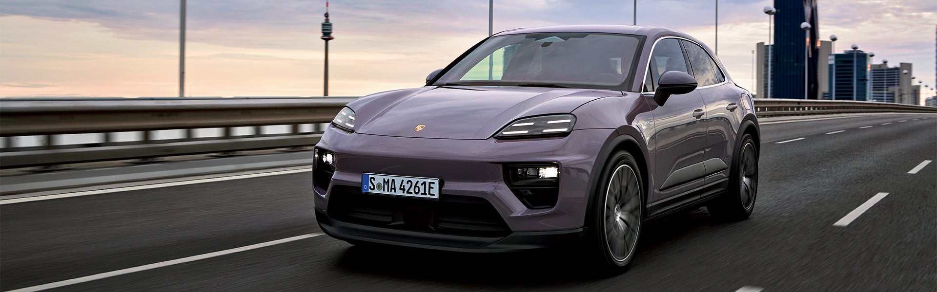 A purple Porsche Macan drives on a highway with blurred city buildings and a tower in the background.