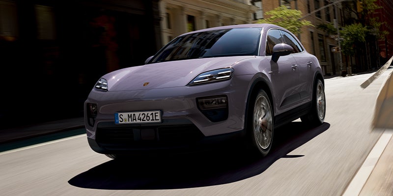 Lavender Porsche Macan EV driving on an urban street with sunlight and dappled shadows.