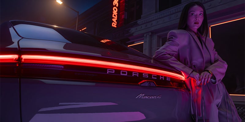 A woman in a stylish suit poses next to the illuminated rear of a Porsche Macan 4 at night.