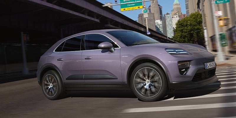 A metallic purple Porsche Macan EV driving on a city street with blurred skyscraper buildings in the background.