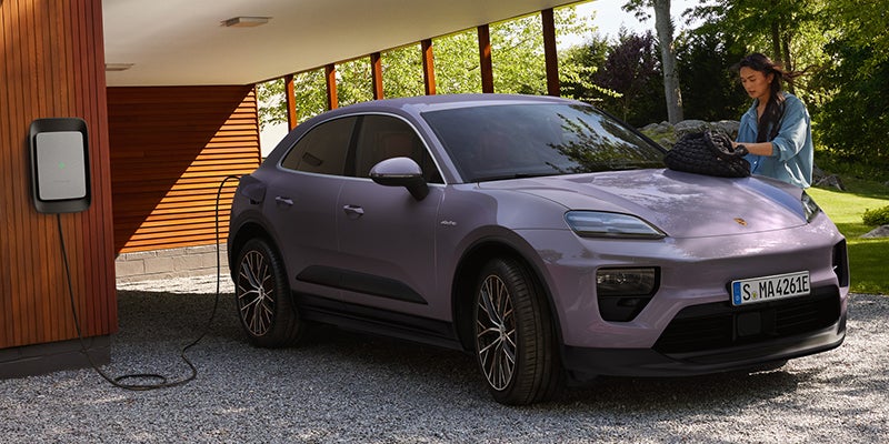 A woman stands beside an electric purple Porsche Macan SUV charging from a home wall charger on a gravel driveway.