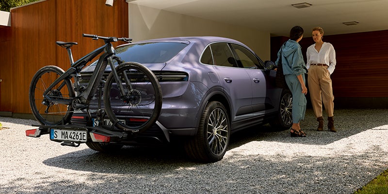 Gray SUV with two bikes on a rear rack, two women chatting beside it in a gravel driveway.