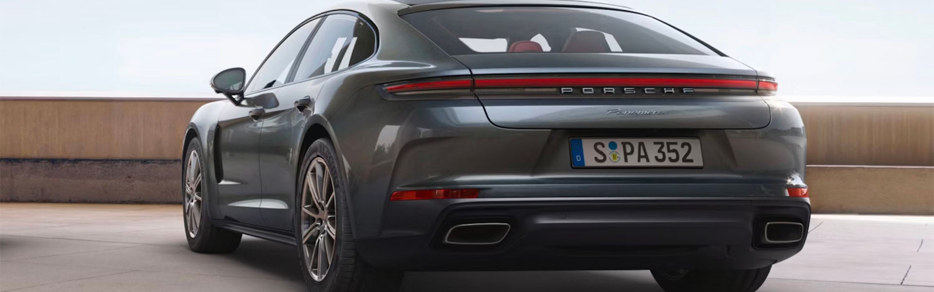Rear view of a dark gray Porsche Panamera parked on a light gray asphalt surface with a beige wall behind.
