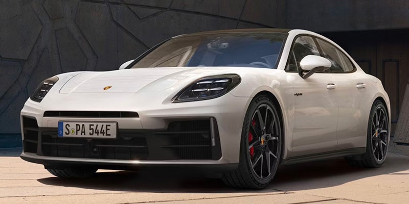 Front 3/4 shot of a white Porsche Panamera parked on a light-colored ground in front of a concrete building.