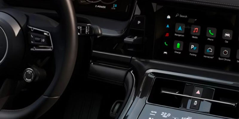 Car interior showing a steering wheel, gear selector, air vent, and a touchscreen display with various app icons.