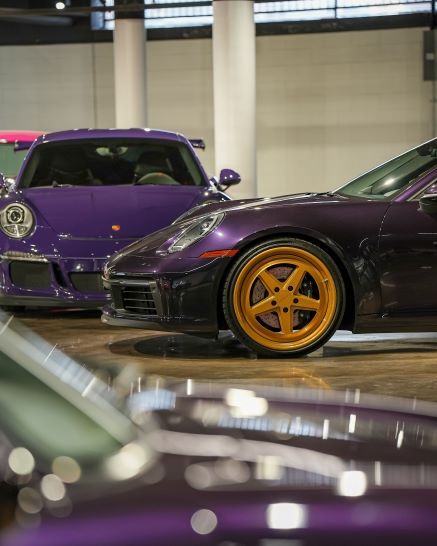 Two purple Porsche sports cars, one with gold wheels, displayed side-by-side in an indoor setting.