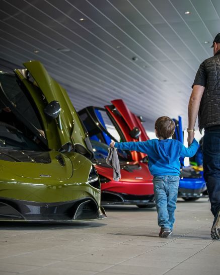A man and a young child in a blue hoodie and jeans walk past several luxury cars with their butterfly doors open.