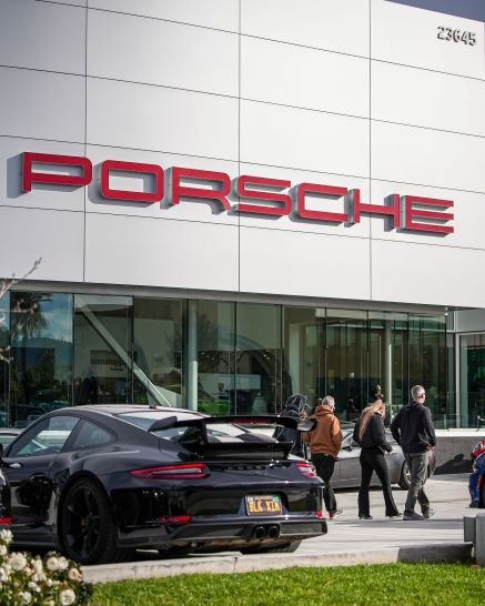 A black Porsche parked in front of a modern dealership with a large red PORSCHE sign and people walking by.