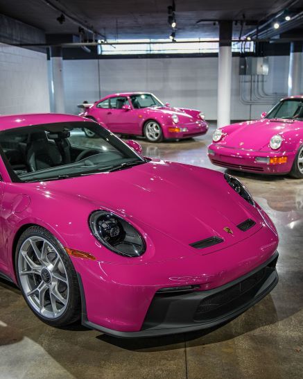 Three hot-pink Porsche sports cars in a garage, with the newest model in the foreground.