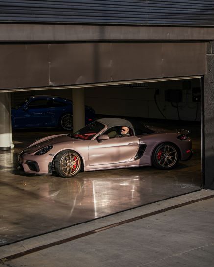 A man with sunglasses driving a champagne-colored Porsche parked in a garage with a blue car behind it.