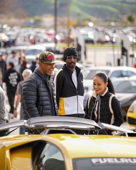 Three people, including Snoop Dogg, laugh while looking at a yellow sports car with an open trunk in a parking lot.