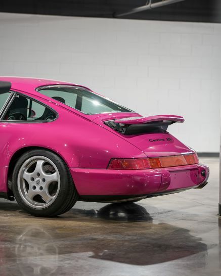 Rear angle of a bright pink Porsche 911 (964) Carrera RS with a spoiler, parked on a glossy concrete floor.