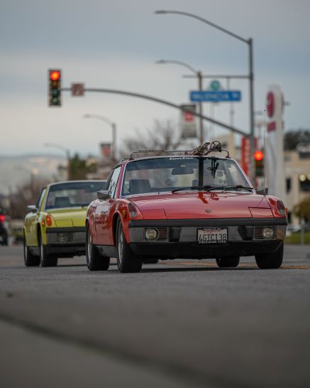 A red vintage Porsche 914 with a surfboard on its roof rack drives on a city street, followed by a yellow 914.