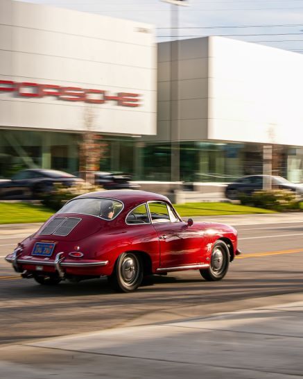 Red vintage Porsche 356 driving past a modern Porsche dealership under a slightly cloudy sky.