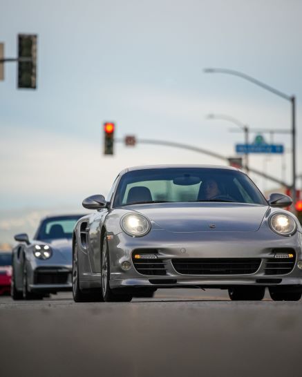 A line of three silver and one red sports cars from the front, with the closest car's headlights on.