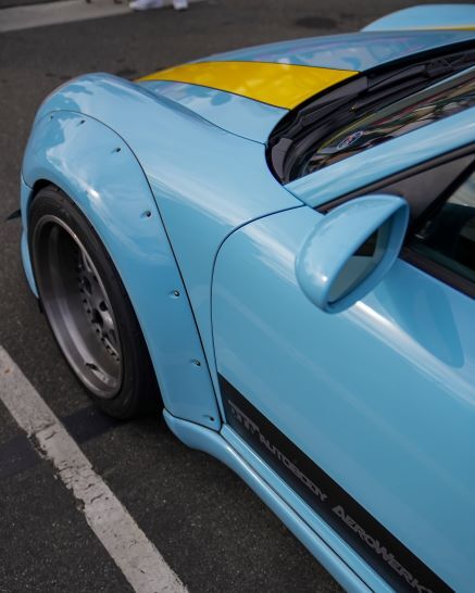 Close-up of a light blue sports car with a yellow racing stripe, wide fender flares, and black side graphics.